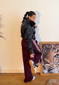 Femme en veste en cuir noire et pantalon de survêtement violet levant la jambe près d'une photo encadrée de léopard sur un sol en bois à l'intérieur.