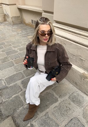 Joven mujer con chaqueta marrón, pantalones blancos y gafas de sol, sentada sobre un pavimento de piedra sosteniendo una taza de café y un bolso negro.
