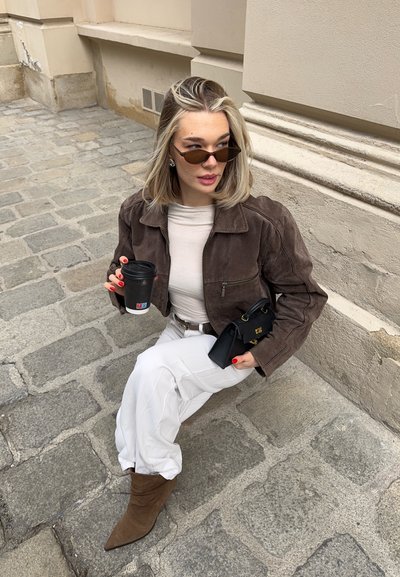 Joven mujer con chaqueta marrón, pantalones blancos y gafas de sol, sentada sobre un pavimento de piedra sosteniendo una taza de café y un bolso negro.