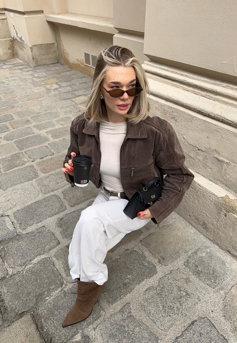 Jeune femme portant une veste marron, un pantalon blanc et des lunettes de soleil, assise sur un pavement en pierre, tenant une tasse de café et un sac noir.