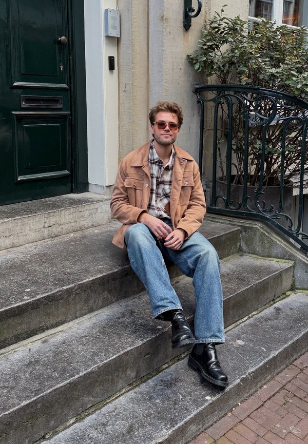 Light brown cord jacket over a checked shirt, blue jeans, and black boots. Seated on stone steps with a green door and wrought iron railing.