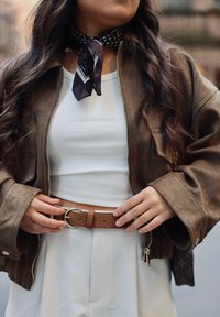 Veste en daim marron, haut blanc côtelé, ceinture en cuir fauve avec boucle argentée, et foulard noir à pois blancs. Pantalon large blanc.