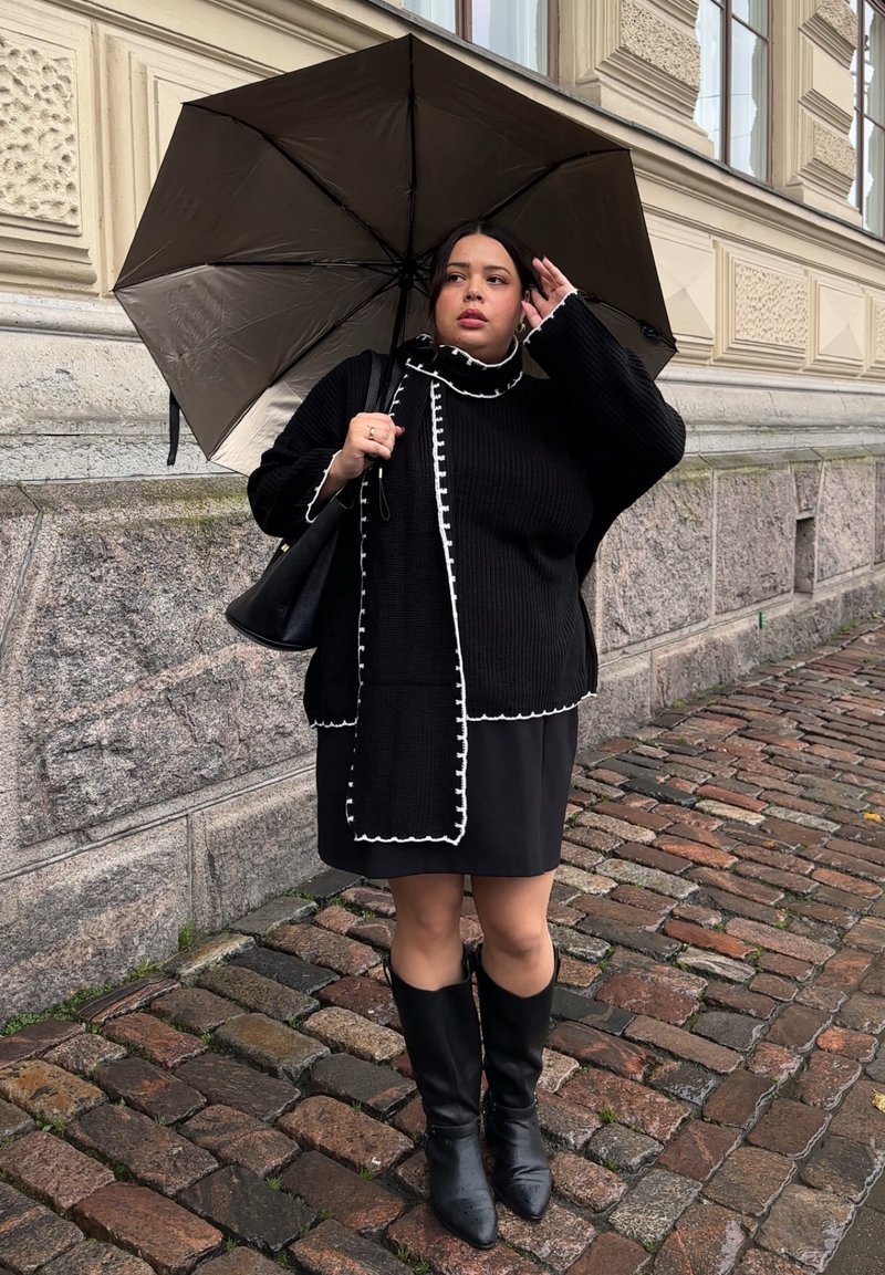 Black knit sweater with white trim, paired with a black skirt and knee-high black boots. Holding a dark umbrella against a stone backdrop.
