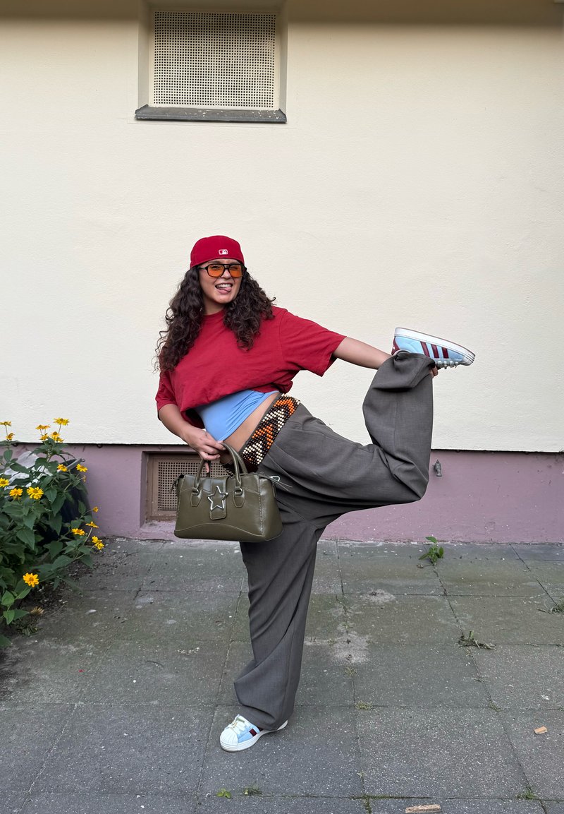 Femme portant une casquette rouge et un haut étire sa jambe derrière en tenant un sac à main vert, debout sur un pied sur un sol pavé près d'un mur avec des fleurs à proximité.