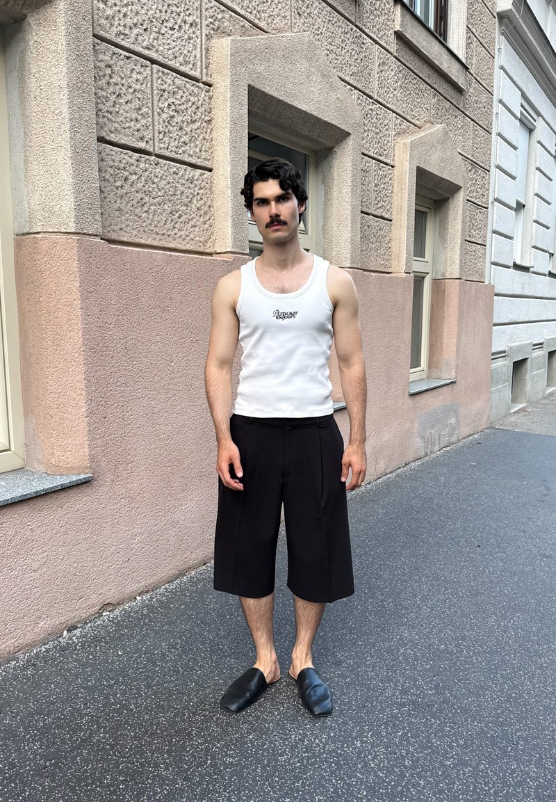 Άνδρας με σκούρα σγουρά μαλλιά και μουστάκι, φορώντας λευκό αμάνικο μπλουζάκι, μαύρα σορτς μέχρι το γόνατο και μαύρα παπούτσια slip-on, στέκεται σε πεζοδρόμιο της πόλης.