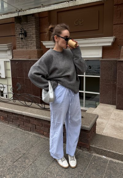 Mujer con suéter gris de gran tamaño y pantalones a rayas bebe café en la calle, lleva gafas de sol y sostiene una bolsa plateada.