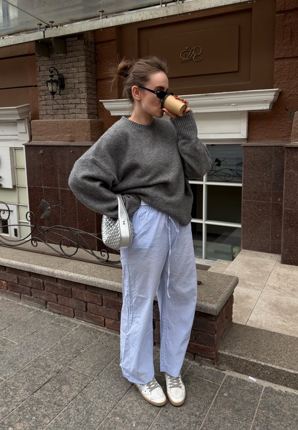 Frau trägt einen oversize grauen Pullover und gestreifte Hose, trinkt Kaffee auf der Straße, trägt eine Sonnenbrille und hält eine silberne Tasche.