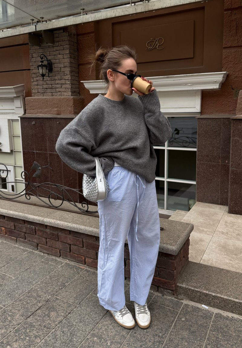 Femme en pull gris oversize et pantalon rayé boit un café dans la rue, porte des lunettes de soleil et tient un sac argenté.