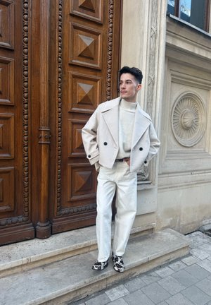Hombre con chaqueta beige, cuello alto blanco y pantalones blancos de pie en escalones de piedra junto a una puerta de madera ornamentada y una pared de piedra tallada.