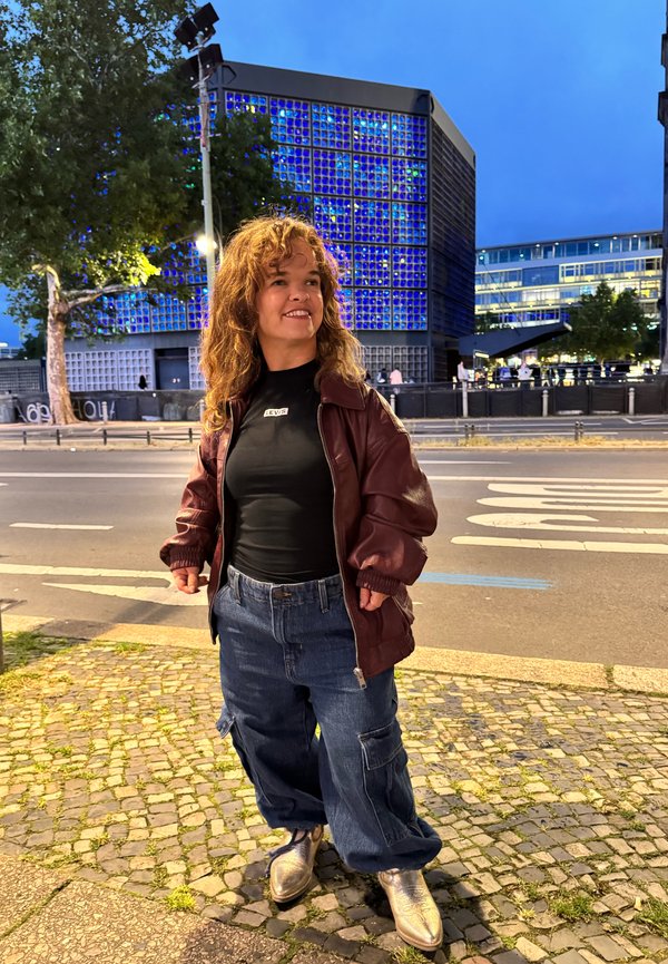 kastanienbraune Lederbomberjacke, schwarzes T-Shirt mit Logo, lockere Jeans-Cargohosen und glänzende silberne Schuhe, stehend auf einem Kopfsteinpflaster an der Straße.