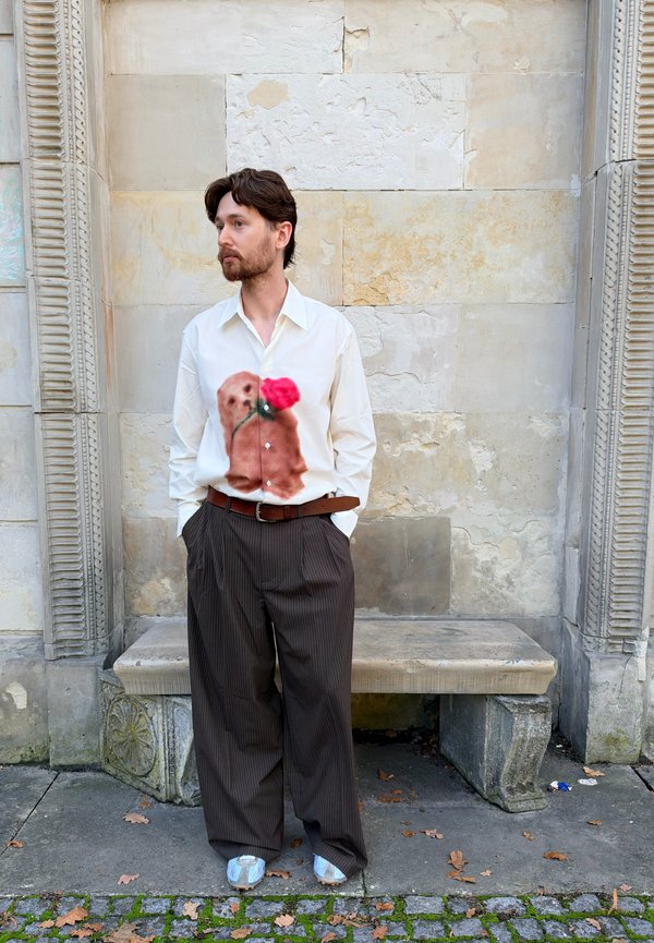 Hombre con cabello castaño y barba que está de pie con las manos en los bolsillos, viste una camisa blanca con estampado de perro y rosa roja, pantalones marrones de rayas y zapatos plateados.