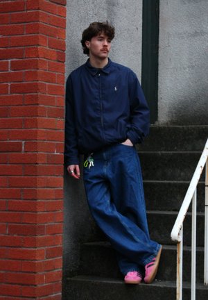 Chaqueta ligera de marina, jeans de denim azul de corte holgado y calzado rosa. De pie contra una pared de ladrillos en unos escalones de concreto.