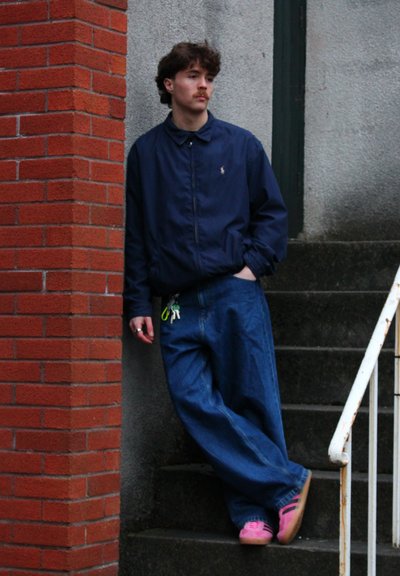 Chaqueta ligera de marina, jeans de denim azul de corte holgado y calzado rosa. De pie contra una pared de ladrillos en unos escalones de concreto.