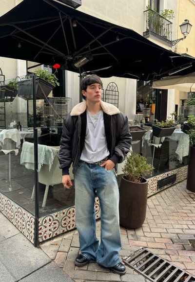 Chaqueta bomber negra con cuello de borrego, camiseta gris y jeans anchos decolorados. Botas negras. Ambiente urbano en una cafetería al aire libre.