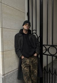 Črna kapuca pod sivim plaščem, kombinirana z rjavimi hlačami z leopardjim vzorcem. Dodatki vključujejo črno beanie klobučko in očala z rumenim odtenkom.
