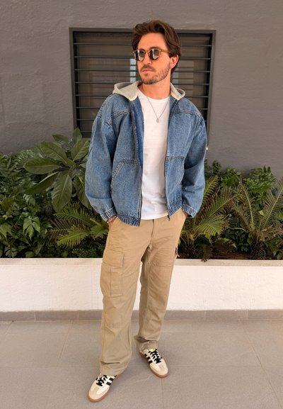 Hombre con gafas de sol, chaqueta vaquera con capucha gris, camiseta blanca, pantalones cargo beige y zapatillas, de pie cerca de plantas contra una pared gris.