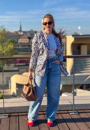 Mujer con blazer de estampado de cebra, top corto blanco, vaqueros anchos, zapatos rojos y gafas de sol, sosteniendo un bolso marrón en una azotea con fondo de paisaje urbano.