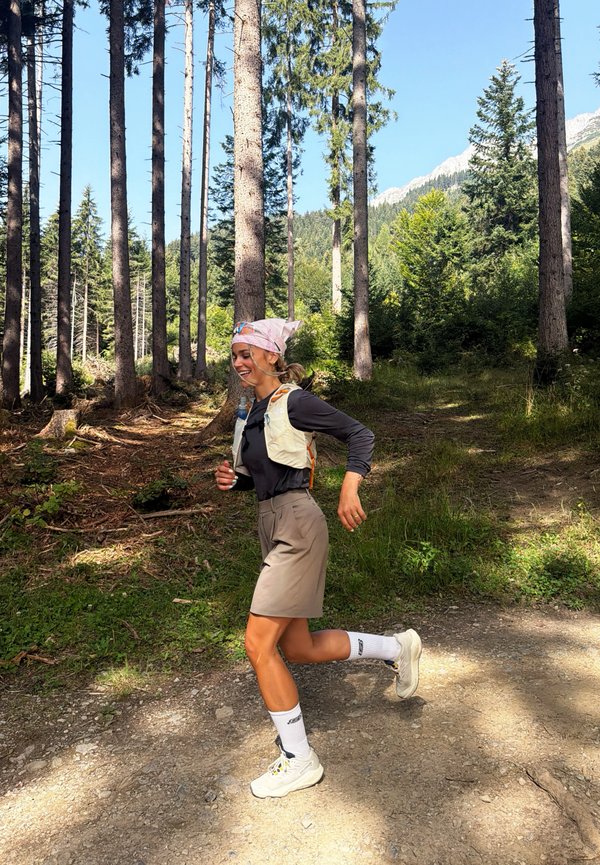Mujer con pañuelo en la cabeza corre por un sendero forestal rodeado de árboles altos y luz solar con montañas visibles en el fondo.