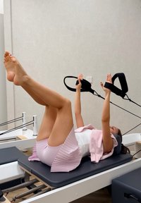 Reformer Pilates-maskin med en svart bas och motståndsremmar; användaren utför benlyft iklädd en vit topp och rosa plisserade shorts.
