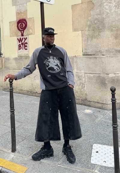 Persona vestida con una camiseta de manga larga negra con acentos grises y un logo, combinada con unas amplias culottes de mezclilla negras y zapatillas negras.