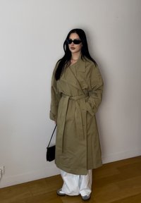 Abrigo trinchera caqui con cinturón, corte oversize y mangas anchas. Combinado con pantalones blancos y accesorios negros, incluyendo gafas de sol y un bolso.