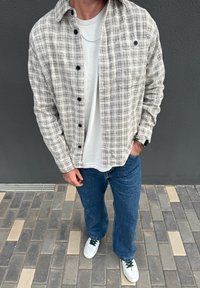 Person wearing a white and black checked button-up shirt over a white T-shirt, blue jeans, white trainers, standing on tiled ground with a grey wall backdrop.