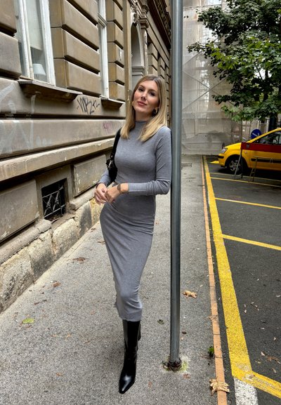 Vestido gris de manga larga ajustado al cuerpo, que llega a medio pantorrilla, combinado con botas negras hasta la rodilla. En el fondo, hay un poste independiente.