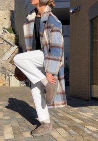 Personne en manteau à carreaux, pantalon blanc et cache-oreilles ajuste des bottines taupe en plein air par une journée ensoleillée avec des escaliers et des bâtiments à proximité.