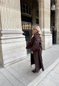 Brun lang kåpe i semsket skinn med belte i midjen, kombinert med en matchende skulderveske og hælede ankelstøvler, plassert foran steinsøyler.