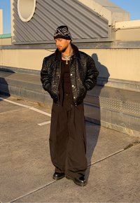 Jachetă bomber neagră matlasată, pulover negru, pantaloni largi negri și bocanci negri. Accesoriile includ coliere argintii suprapuse și o căciulă cu imprimeu.