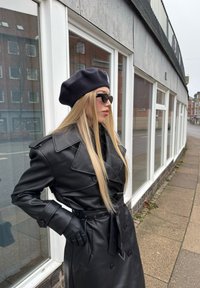 Trench-coat en cuir noir avec de larges revers, taille ceinturée et détails à boutons. Accessoires comprenant un béret noir et des lunettes de soleil foncées.