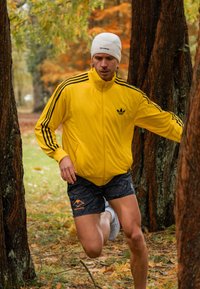 Biegający mężczyzna w jasnoniebieskiej rozpinanej kurtce z trzema czarnymi paskami, czarnych szortach w wzory i białej czapce beanie, otoczony drzewami.