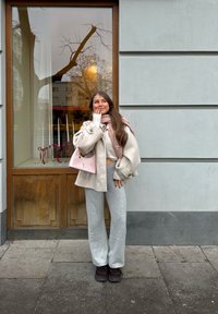 Jeune femme vêtue d'un manteau beige et d'un pantalon gris se tenant sur le trottoir, posant avec la main près de la bouche devant une fenêtre en bois affichant des chandeliers ornés de rubans rouges.