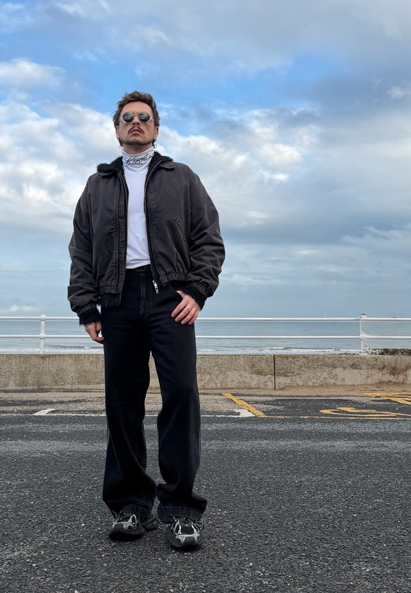 Un homme portant des lunettes de soleil, une veste noire et un pantalon noir se tient sur une promenade en bord de mer sous un ciel nuageux.