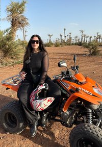 Veicolo tutto terreno (ATV) arancione con accenti neri, pilota in abbigliamento nero, occhiali da sole, che tiene un casco rosso e bianco. Sfondo desertico.