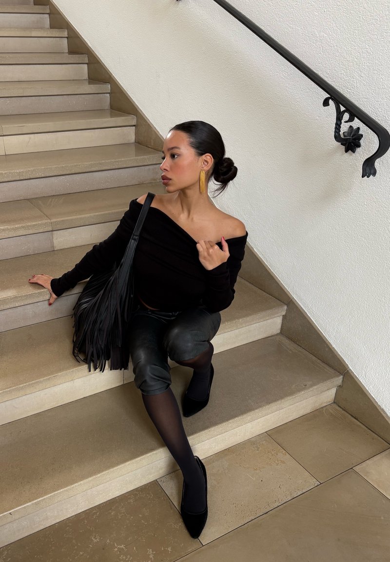 Młoda kobieta w czarnym topie bez ramion, skórzanych spodniach i torbie z frędzlami siedzi na wewnętrznych kamiennych schodach, patrząc w bok.