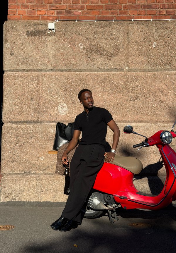 Hombre vestido de negro apoyado en un scooter rojo estacionado junto a una pared de piedra texturizada bajo la luz del sol.
