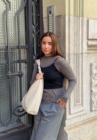 Schwarzes ärmelloses Oberteil mit grauen langen Ärmeln, kombiniert mit hochtaillierten grauen Hosen und einer cremefarbenen, strukturierten Schultertasche, vor einer kunstvoll verzierten Tür.