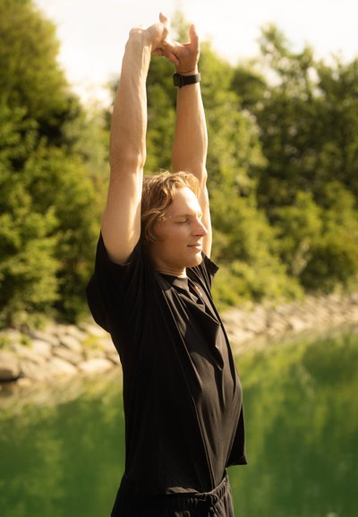 Camisa y pantalones cortos deportivos negros. Persona estira los brazos por encima de la cabeza, con los ojos cerrados. Agua verde y árboles de fondo. Luz natural suave.