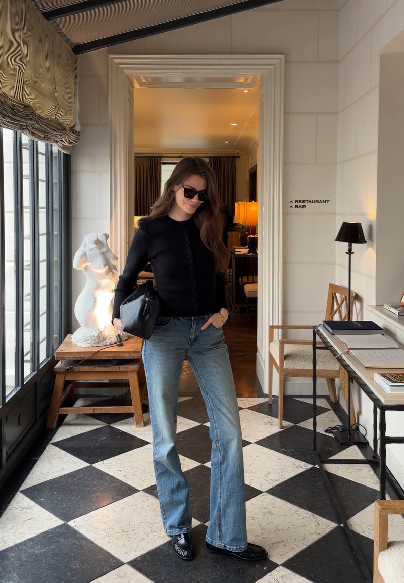 Femme en haut noir, lunettes de soleil et jeans tenant un sac, debout sur un sol carrelé noir et blanc près d'une sculpture et d'un panneau de restaurant.