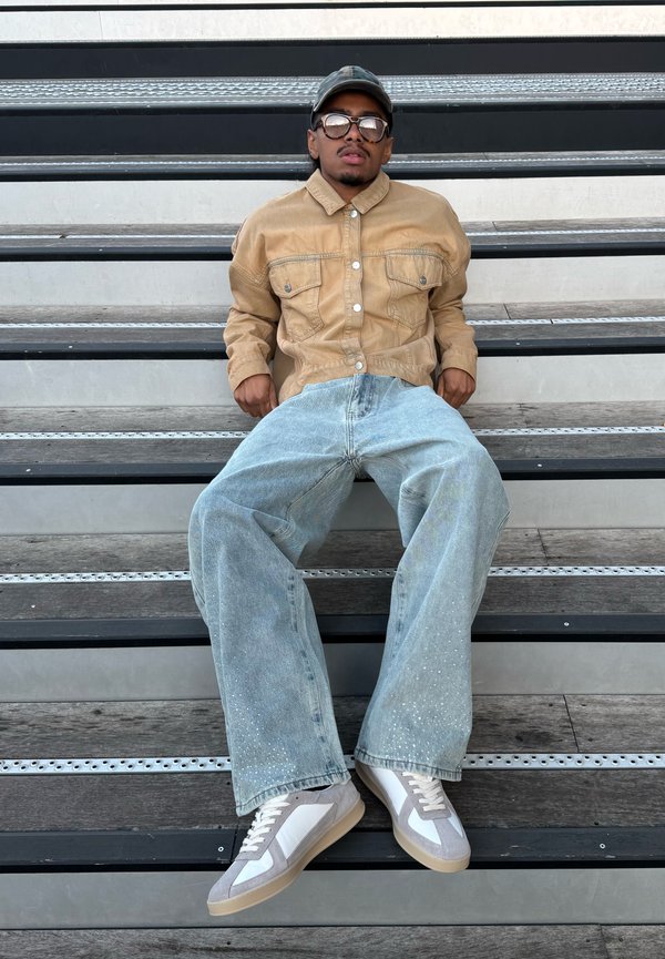 Joven con chaqueta marrón claro, vaqueros anchos azul claro, zapatillas, gorra y gafas de sol sentado en gradas de metal y madera.