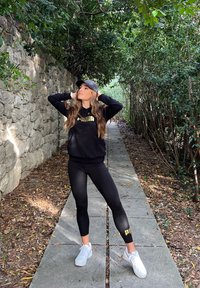 Sudadera negra con capucha y logo dorado, leggings negros ajustados con el logo de la marca y zapatillas blancas. La modelo posa en un camino de piedra rodeado de vegetación.
