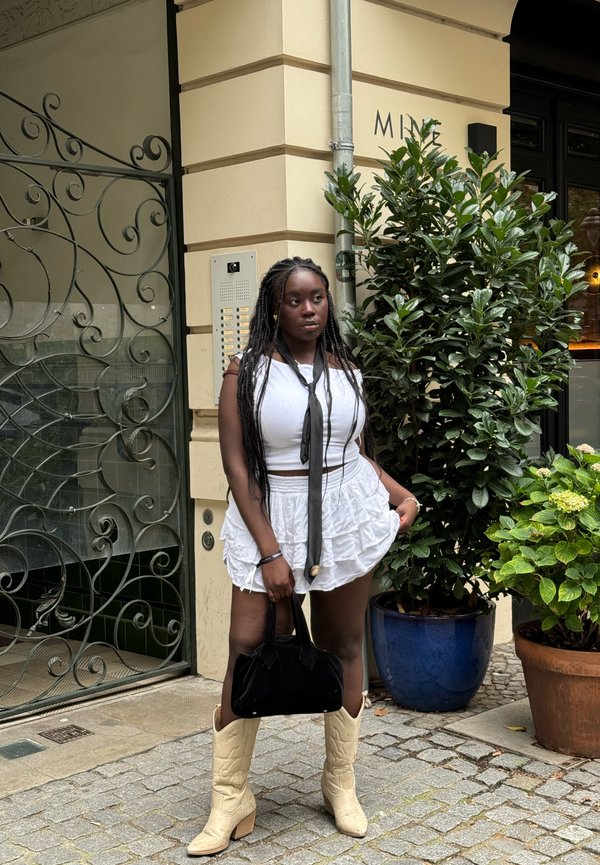 Gonna bianco con rouches, top bianco senza spalline, cravatta nera e stivali da cowboy beige. Tiene una borsa nera. Lo sfondo include piante e architettura.