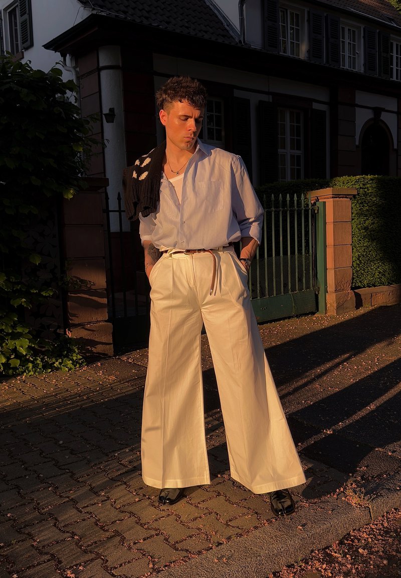 Person trägt weite beige Hosen, hellblaues Hemd und schwarze Schuhe und steht auf einem sonnigen Gehweg vor einem Haus mit grünem Tor.