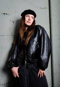 Μαύρο δερμάτινο μπόμπερ με ψηλό γιακά, ριγωτά μανίκια και ζώνη; συνδυασμένο με μαύρη δαντελένια μπλούζα και σκούρους ψηλόμεσους τζιν.