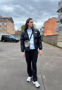 Čierna bunda s kapucňou, svetlomodrý pruhovaný sveter, čierne teplákové nohavice, sivá crossbody taška a biele tenisky v vonkajšom prostredí.