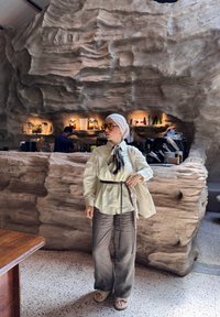 Donna con velo e occhiali da sole gialli si trova vicino a un bancone di caffè dalla texture simile a una caverna, uomo che lavora dietro il bar con attrezzature per caffè.