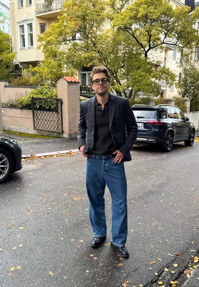 Blazer negro sobre una camisa oscura texturizada, combinado con jeans anchos azules y zapatos negros. Ambiente al aire libre con hojas de otoño en el pavimento.