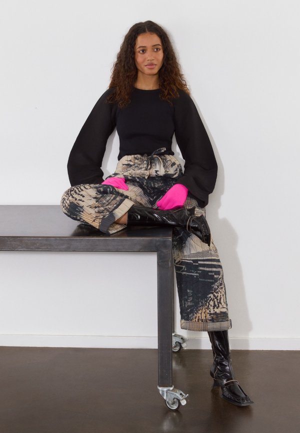 Top negro de manga larga, pantalones de pierna ancha de estampado en beige y negro, guantes vibrantes en color rosa, y botas negras hasta la rodilla, sentado sobre una mesa.