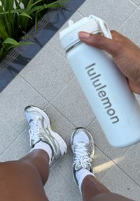 Personne portant des chaussures de course blanches et argentées et des chaussettes blanches, tenant une bouteille d'eau blanche Lululemon, debout sur un sol carrelé gris près de plantes vertes.